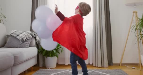 Bambino in costume da supereroe con mantello rosso e maschera che gioca con palloncini in un ambiente domestico. Casual concetto di tempo libero per interni. Filmati 4k di alta qualità - Filmati, video