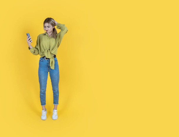 Irritated confused young woman, full body length front view of irritated confused young woman. Hold use smartphone on hand looking screen scratching her head. Reading bad news. Yellow background. - Photo, Image