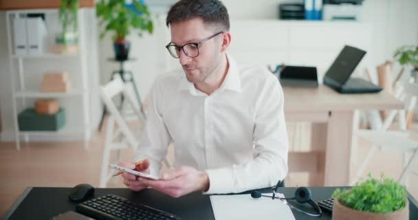 Fiducioso imprenditore maschio scorrimento internet su tablet digitale mentre seduto alla scrivania con auricolare presso lo spazio di lavoro aziendale - Filmati, video