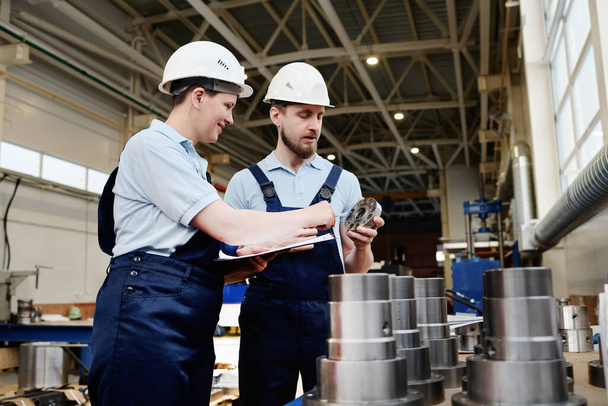 Średnie ujęcie mężczyzny i kobiety w mundurach i twardych kapeluszach pracujących w fabryce, kontrolujących jakość. - Zdjęcie, obraz