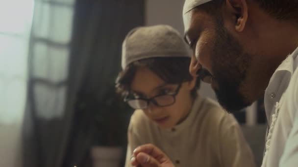 Сторона крупным планом Biracial бородатый отец в традиционной одежде чтение мини Коран книги и объяснение исламской религии своему маленькому сыну дома - Кадры, видео
