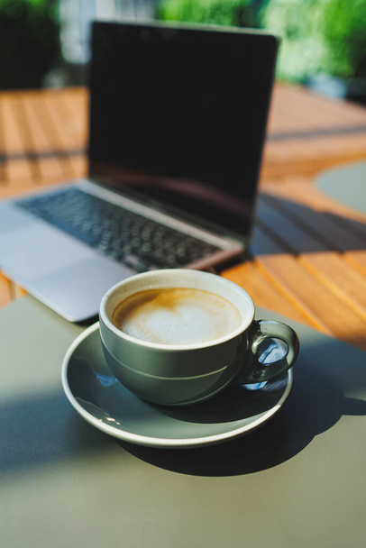 Yazlık terasta dizüstü bilgisayarın arka planında bir fincan aromatik kahve. Bir kafede çalışıyorum. Uzaktan çalışma çevrimiçi. - Fotoğraf, Görsel