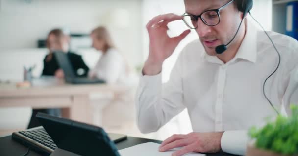 Zelfverzekerde zakenman die via een koptelefoon praat terwijl hij op papier berekent en schrijft op de werkplek van het bedrijf - Video