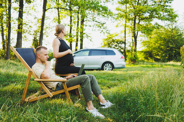Ein Familienpaar, ein Mann und eine Frau, entspannen sich in der Natur und arbeiten aus der Ferne mit einem Laptop. Picknick liebevoller Paare mit Online-Arbeit. Eine schwangere Frau ruht mit ihrem Mann in der Natur - Foto, Bild
