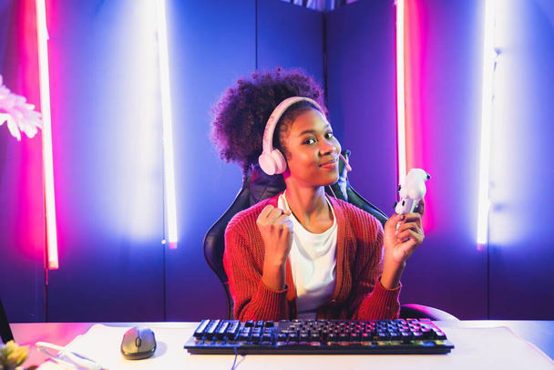 Победительница афроамериканской женской игровой команды, играющая в онлайн-боях с Esport, носит наушники в комнате неонового освещения. Говоря другие игроки планируют стратегии, чтобы выиграть конкурентов. Tastemaker. - Фото, изображение