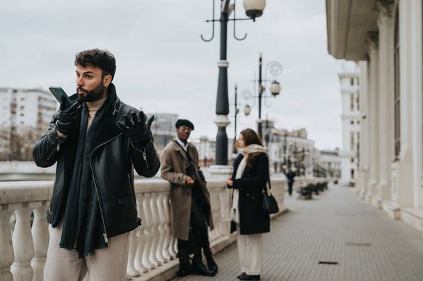 Un joven hombre de negocios barbudo con una chaqueta de cuero está teniendo una conversación telefónica en una calle de la ciudad, con gente caminando por el fondo. - Foto, Imagen