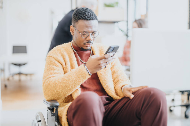 Ένας προσεκτικός νεαρός σε αναπηρική καρέκλα ασχολείται με το έξυπνο τηλέφωνό του σε ένα σύγχρονο περιβάλλον γραφείου, παραδειγματίζοντας την προσβασιμότητα και την ένταξη. - Φωτογραφία, εικόνα