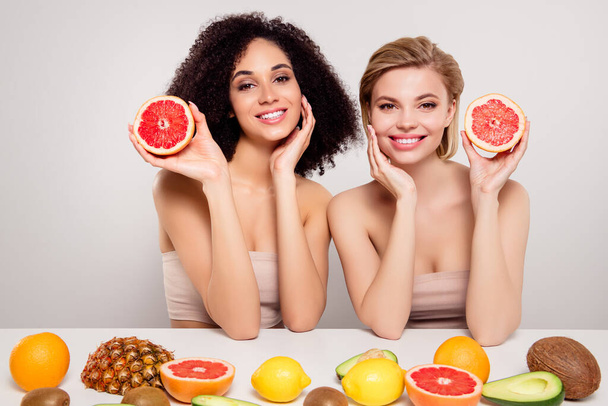 Фото великолепных девушек сидят стол держать половину ломтик грейпфрута прикосновение идеальной кожи лица на пастельном фоне цвета. - Фото, изображение