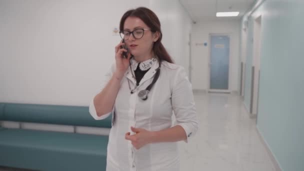 Young woman dotkor walks down the corridor of hospital and talks on cell phone. Female physician makes phone call passes through the clinic hallway. Healthcare professional talking to patient.  - Footage, Video