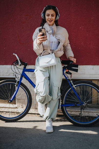 Modna młoda kobieta słucha muzyki na słuchawkach i sprawdza swój smartfon stojąc przy rowerze w słoneczny dzień. - Zdjęcie, obraz