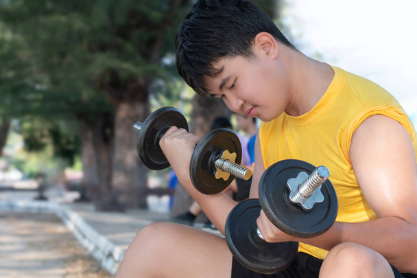 Ázsiai kövér fiatalember emel négy kilogramm és nyolc kilogramm súlyzók gyakorolni és lefogy egy nyitott udvaron késő délután, egészségügyi ellátás és szabadidős tevékenység koncepció. - Fotó, kép