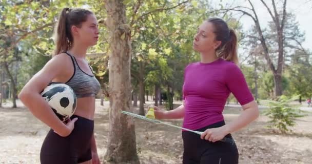 Twee gefocuste vrouwelijke atleten in sportkleding met een voetbal en een badmintonracket in een parksetting die een gesprek voeren terwijl ze even uitrusten. - Video