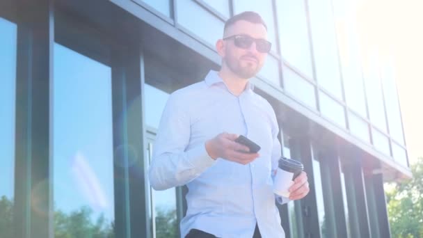 Een man met een zonnebril en een baard staat voor een gebouw en kijkt naar zijn mobiele telefoon. Bedrijfsconcept - Video