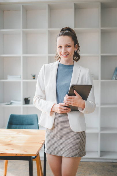 Business-Management-Konzept: Eine lächelnde junge Frau steht in einem modernen Büro und hält ein Tablet in der Hand, bereit für einen produktiven Arbeitstag. - Foto, Bild