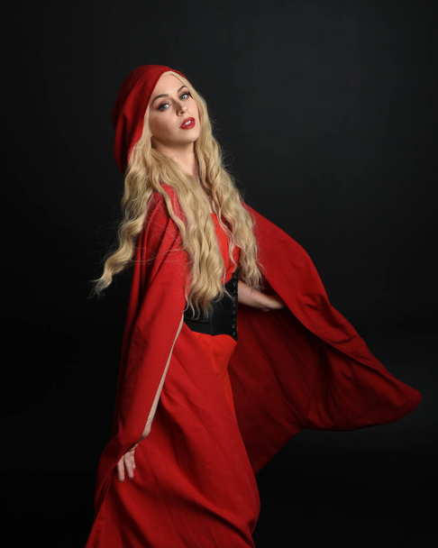 zbliżenie portret piękny model z długimi blond włosy noszenie efektowny strój fantasy z gorsetem i płynące czerwone kaptur bajkowy płaszcz jazdy. izolowany na ciemnym tle studio. - Zdjęcie, obraz
