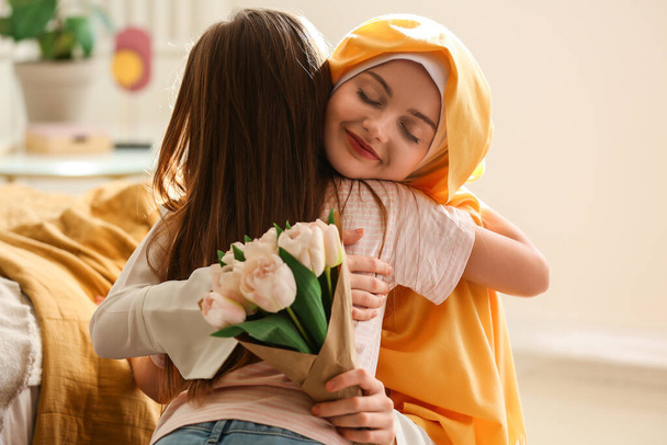 Mujer musulmana feliz con tulipanes abrazando a su hija pequeña en casa en el Día de la Madre - Foto, Imagen