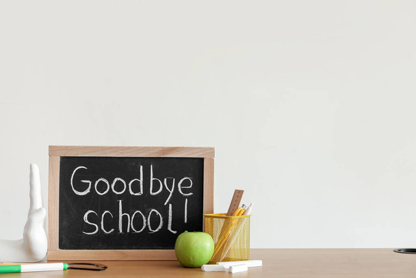 Krijtbord met tekst GOODBYE SCHOOL, appel en briefpapier op tafel bij lichtmuur - Foto, afbeelding