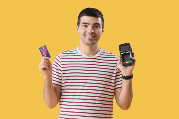 Junger Mann mit Zahlungsterminal und Kreditkarte auf gelbem Hintergrund - Foto, Bild