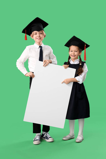 Niños pequeños con sombreros de graduación con póster en blanco sobre fondo verde. Fin del año escolar - Foto, imagen