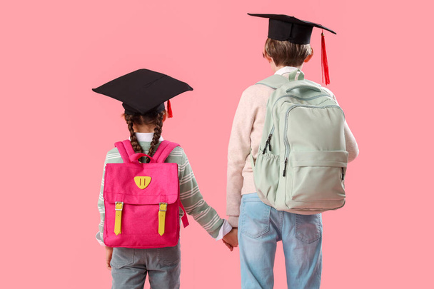 Petits enfants dans des chapeaux de graduation avec des sacs à dos sur fond rose, vue arrière. Fin d'année scolaire - Photo, image