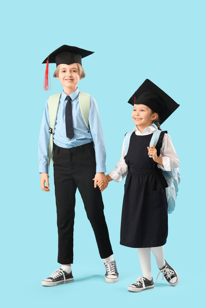 Niños pequeños en sombreros de graduación con mochilas caminando sobre fondo azul. Fin del año escolar - Foto, imagen