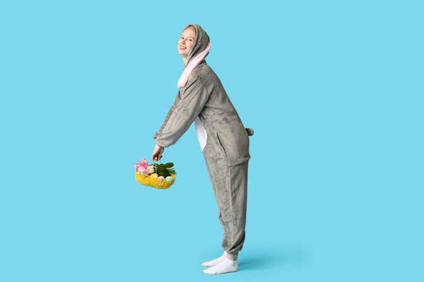 Красива молода щаслива жінка в костюмі кролика тримає кошик з яйцями і букет тюльпанів на синьому фоні. Великоднє святкування - Фото, зображення