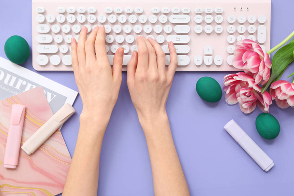 Γυναικεία χέρια με πληκτρολόγιο υπολογιστή, πασχαλινά αυγά, σετ γραφικής ύλης, λουλουδιών και περιοδικού σε λιλά φόντο - Φωτογραφία, εικόνα
