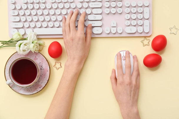 Female hands with Easter eggs, flowers, cup of tea, computer keyboard and mouse on yellow background - Photo, Image