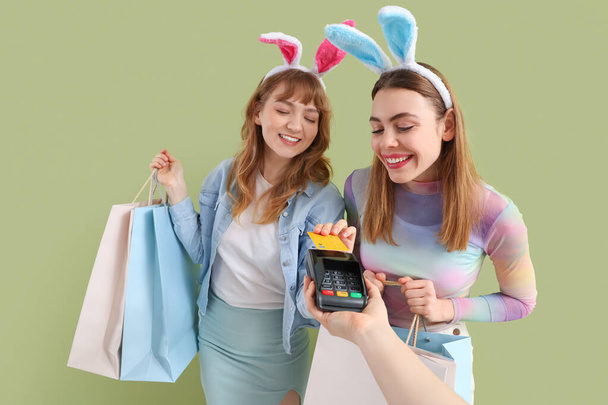 Felice sorridente giovani donne in orecchie di coniglio fasce in possesso di carta di credito, borse della spesa di carta per Pasqua e mano con terminale di pagamento su sfondo verde - Foto, immagini
