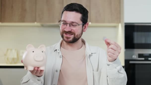 Portrait of intelligent male with beard weighting piggy bank and coin in hands while standing at kitchen. Doubting man thinking about making saving or wasting money and looking at different options. - Footage, Video