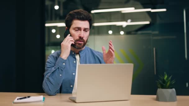 Soustředěný muž s vousy mluví po telefonu a gestikuluje rukama. Mužská podpora agent poradenství klientů a pomáhá řešit technický problém s internetem při sezení na pracovišti s notebookem. - Záběry, video