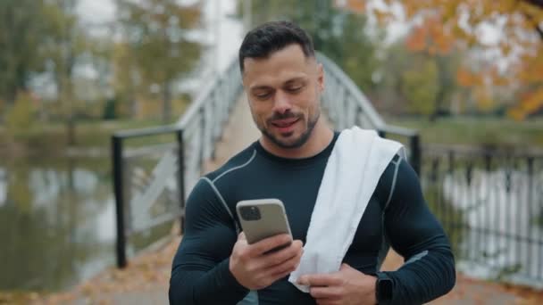 sportif beau mature caucasien homme marcher dans l 'parc avec serviette sur son épaule et en utilisant smartphone texto ou calculer les calories après son extérieur courir - Séquence, vidéo