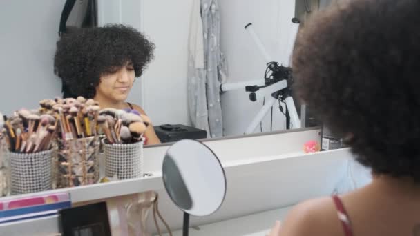 Trans γυναίκα χρησιμοποιώντας το κινητό τηλέφωνο πριν αρχίσει να συγκεντρώνει τα μαλλιά της μπροστά από έναν καθρέφτη στο σπίτι - Πλάνα, βίντεο