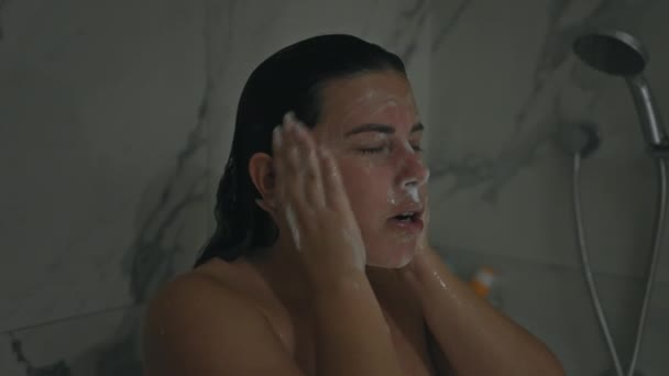 Una mujer joven aplica un limpiador facial en un baño de baldosas de mármol, que representa la higiene y el cuidado. - Imágenes, Vídeo