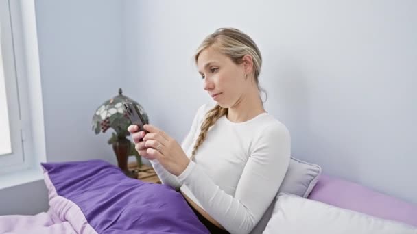 Una giovane donna bionda a casa usando il suo smartphone mentre riposa a letto con il suo cane labrador nero. - Filmati, video