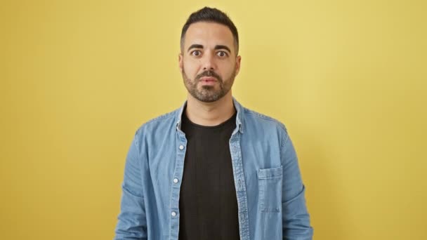 Ударний молодий іспаномовний чоловік в джинсовій сорочці, що стоїть з подивом і невірою, виразне обличчя наповнене шоком і страхом на ізольованому жовтому тлі - Кадри, відео