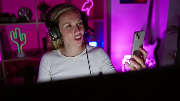 Μια νεαρή ξανθιά γυναίκα συμμετέχει σε μια βιντεοκλήση σε ένα δωμάτιο με νέον φως τη νύχτα, απεικονίζοντας συνδεσιμότητα και αναψυχή σε εσωτερικούς χώρους. - Πλάνα, βίντεο