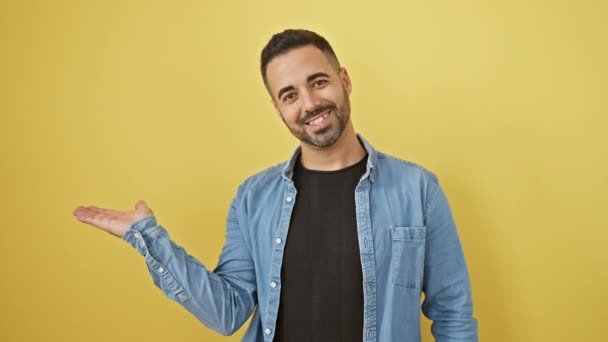 Iloinen nuori latinomies denim-paidassa, joka esittelee tervetuliaiseleen, hymyilee ja osoittaa kämmenellä keltaisella eristetyllä taustalla - Materiaali, video