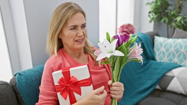 Ανίδεη μεσήλικη ξανθιά γυναίκα στο σπίτι, χωρίς αμφιβολία κρατώντας δώρο και λουλούδι μπουκέτο με προβληματισμένη έκφραση - Πλάνα, βίντεο