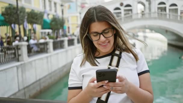 Giovane donna utilizzando smartphone dal canale in stile veneziano in doha, occhiali chic, sms, viaggi, all'aperto. - Filmati, video