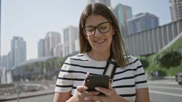 Μια νεαρή γυναίκα χρησιμοποιεί ένα smartphone σε μια ηλιόλουστη οδό Ντουμπάι με ουρανοξύστες στο παρασκήνιο. - Πλάνα, βίντεο