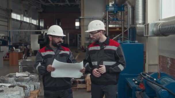 Střední snímek dvou etnicky různorodých mužských inženýrů v pracovních oděvech a tvrdých kloboucích diskutujících o modrotiscích a sledujících detaily hardwaru při společné práci v průmyslových závodech - Záběry, video