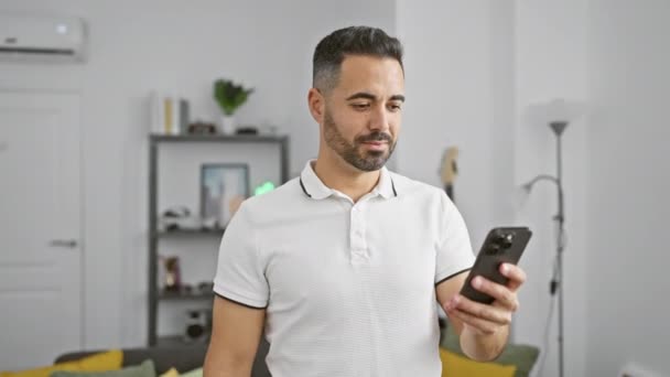 Счастливый, позитивный молодой латиноамериканец, подающий знак одобрения дома, уверенно улыбающийся со смартфоном, чувствующий себя успешным парнем - Кадры, видео