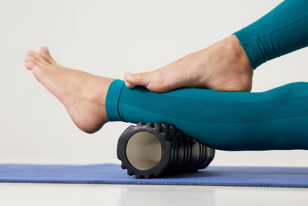 девушка массирует ноги на ролике и делает йогу на белом фоне, массаж ролика для фитнеса и йоги, домашняя тренировка - Фото, изображение