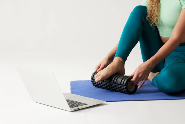 niña sosteniendo un rodillo de masaje mientras está sentado en el suelo y cerca de un ordenador portátil, rodillo de masaje para la aptitud y el yoga, entrenamientos en línea en casa - Foto, Imagen