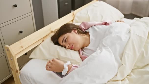 Młoda kobieta korzysta ze smartfona, relaksując się w łóżku w przytulnych pomieszczeniach swojej sypialni. - Materiał filmowy, wideo