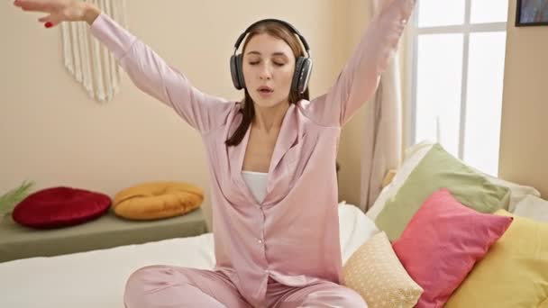 Μια νεαρή γυναίκα που απολαμβάνει μουσική με ακουστικά σε ένα άνετο σκηνικό κρεβατοκάμαρας, φορώντας ένα ροζ σετ πιτζάμας. - Πλάνα, βίντεο