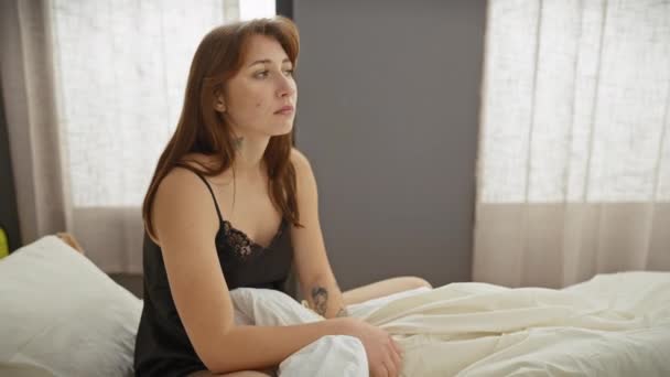 Eine nachdenkliche junge Frau sitzt in einem Schlafzimmer, durch Vorhänge dringt Sonnenlicht.. - Filmmaterial, Video