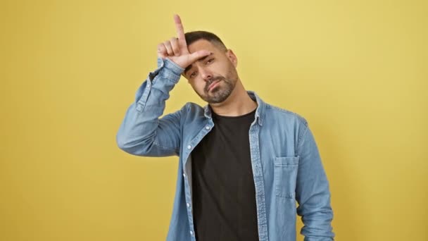 Frecher junger hispanischer Mann im Jeanshemd, vor gelbem Hintergrund stehend, beleidigt Menschen mit Loser-Geste - Finger an die Stirn, spottet und lacht unhöflich - Filmmaterial, Video