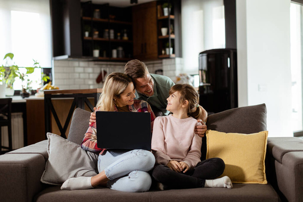 Iloinen kolmen hengen perhe viettää laatuaikaa yhdessä olohuoneen sohvalla, jakaen hetken kannettavan tietokoneen ympärillä mukavassa kodissaan. - Valokuva, kuva